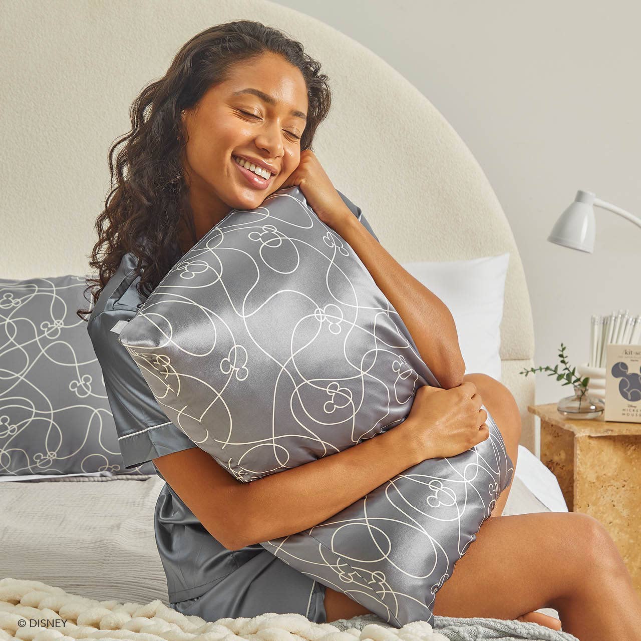 Blyskt Woman in a gray satin robe holding a matching pillow with a floral pattern in a bedroom setting.