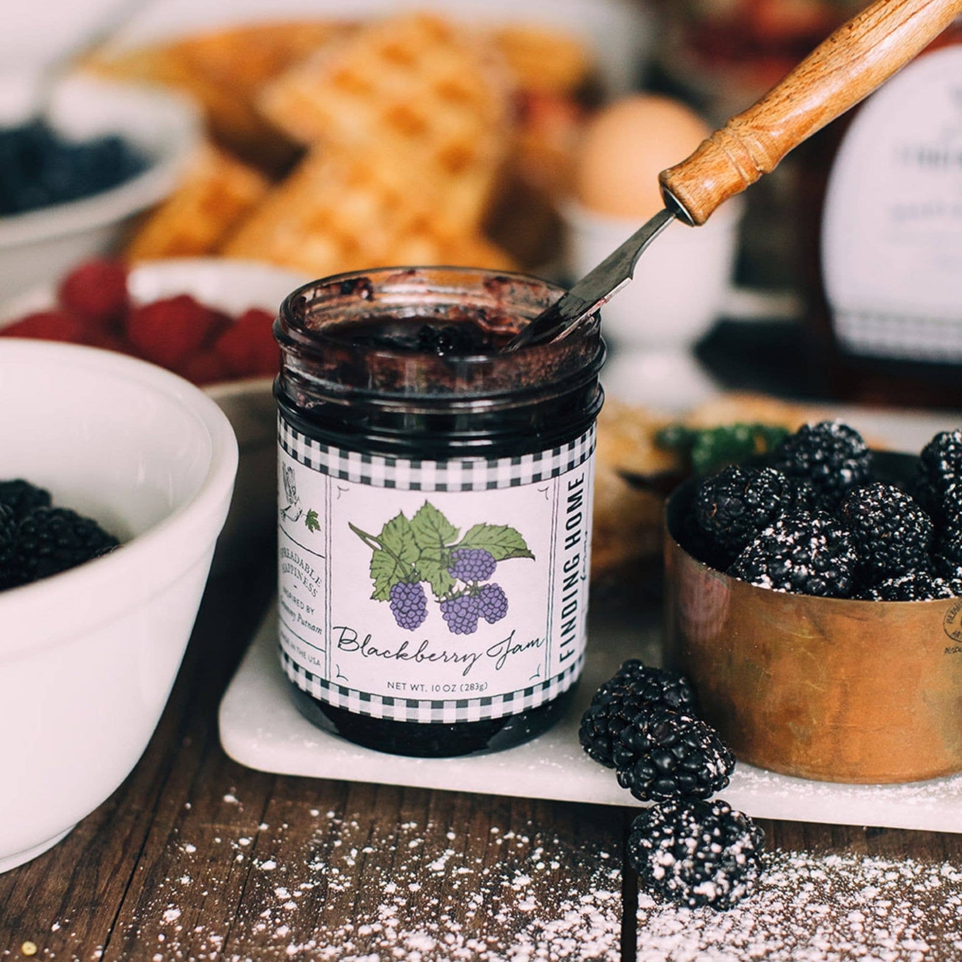 Blyskt Jar of blackberry jam with a spoon, surrounded by blackberries and pastries on a wooden table.