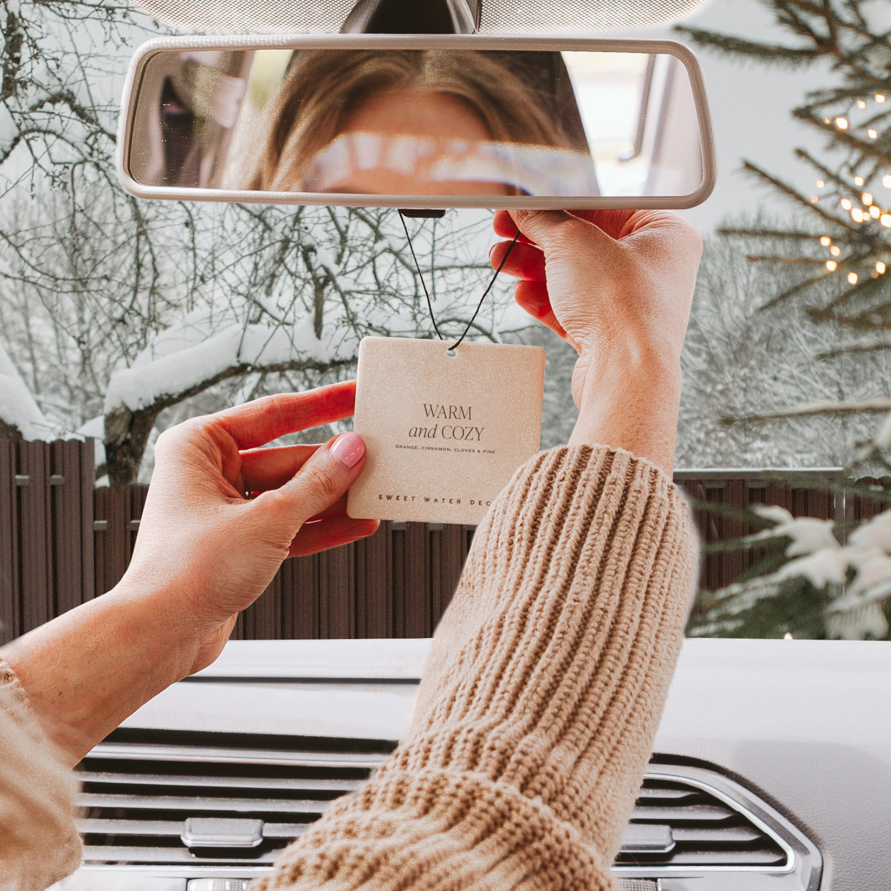 Person holding a card with 'Warm and Cozy' text in front of a car's rearview mirror on a snowy day.