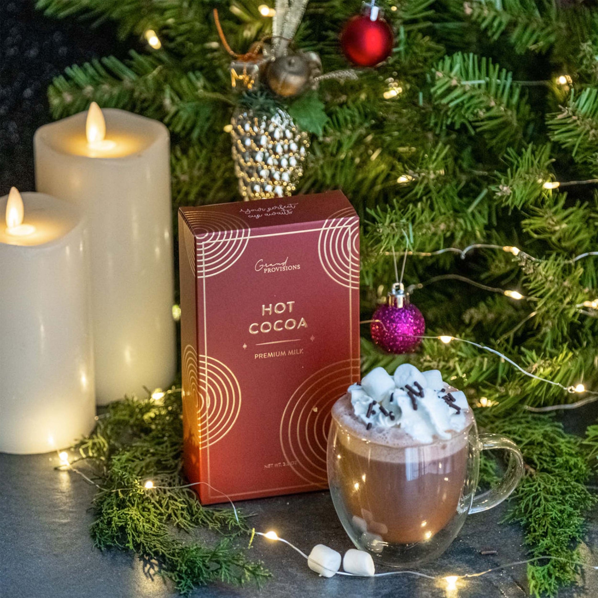 Blyskt Hot chocolate with marshmallows, a box of hot cocoa, and candles in front of a Christmas tree.